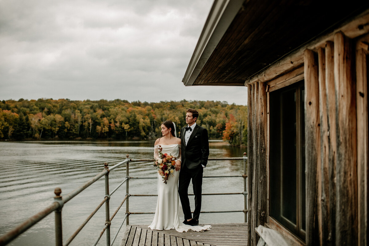 Newlyweds wearing chic wedding attire gaze over a peaceful lake amongst vibrant fall foliage.