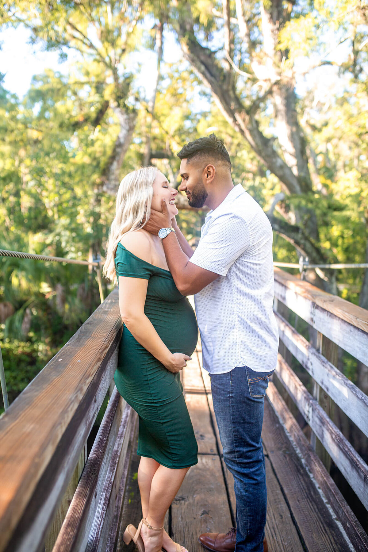 Dark haired man holds woman in green dress face while bringing her in for a kiss standing on a bridge 