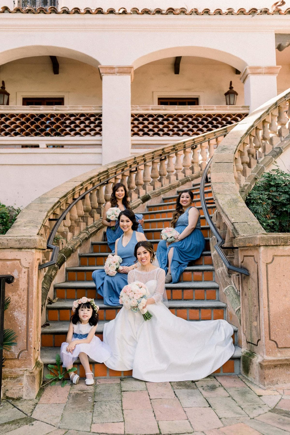 Bride sits on a spiral staircase with her dress train fanned out and the bridesmaids sitting all around her