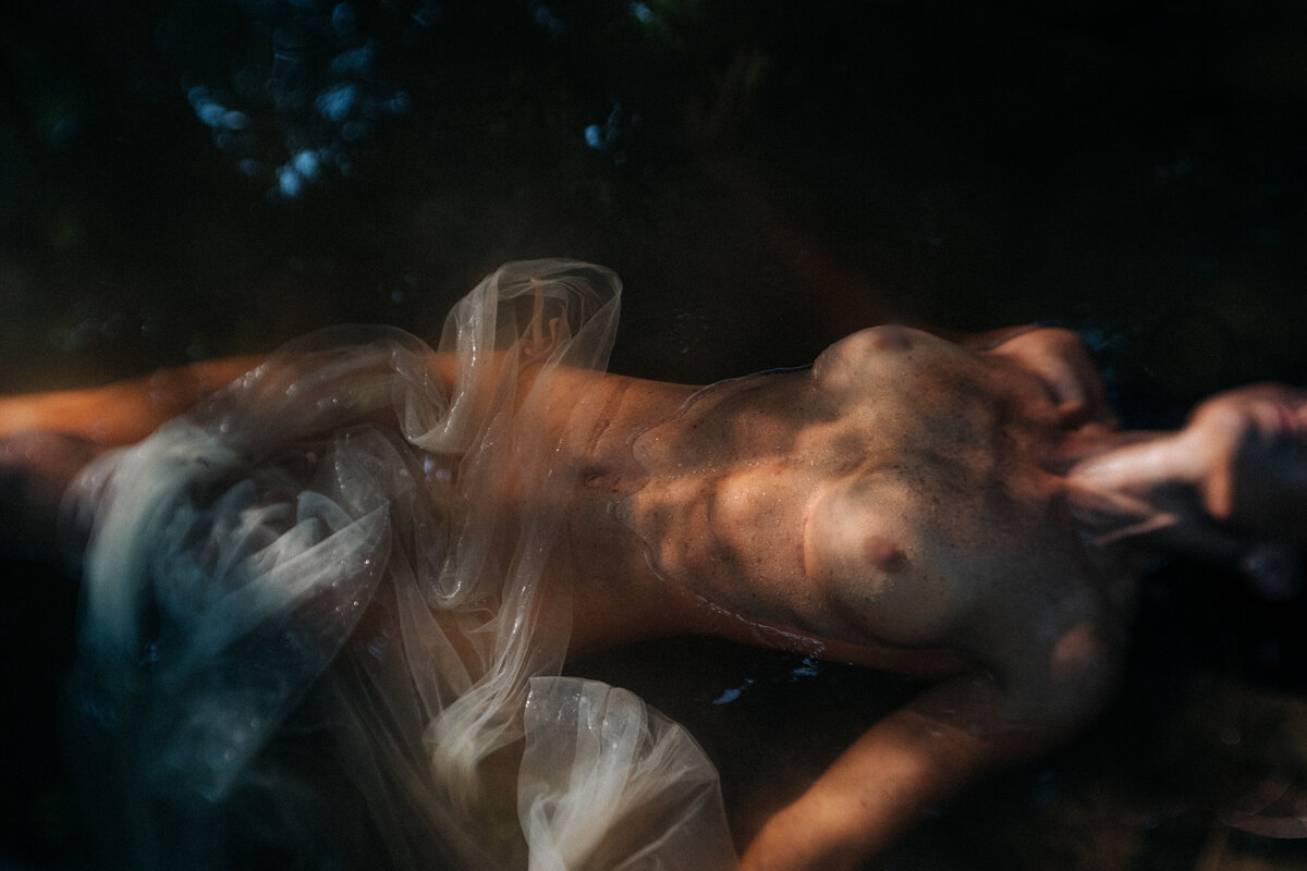 A partially covered, bare-chested person lies in water with dappled light and shadows on their skin. Translucent fabric is draped over the lower part of their body. Fine Art Boudoir Photography, Durham NC