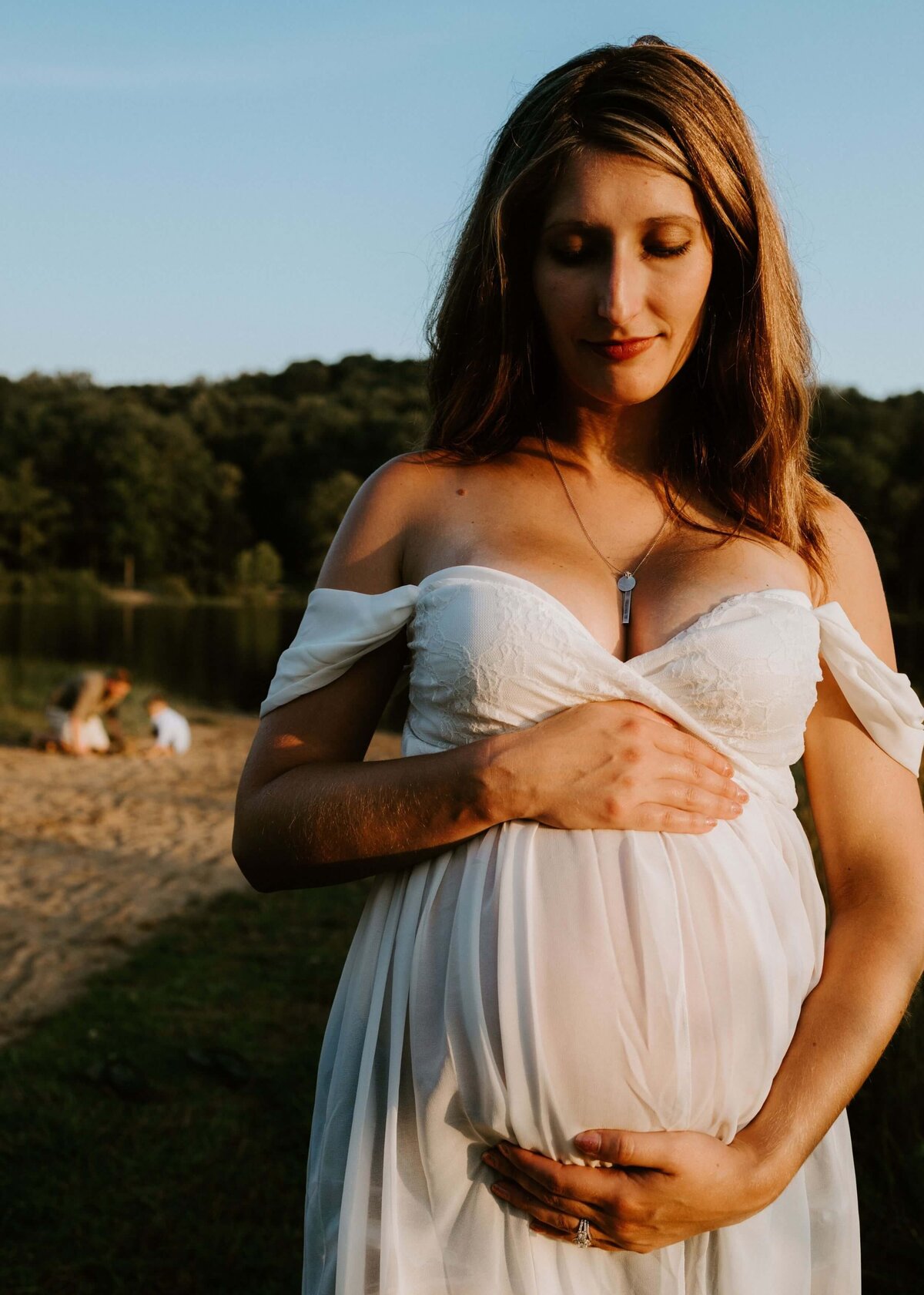 A pregnant woman in a white dress elegantly poses by a serene lake, captured by a Pittsburgh maternity photographer.