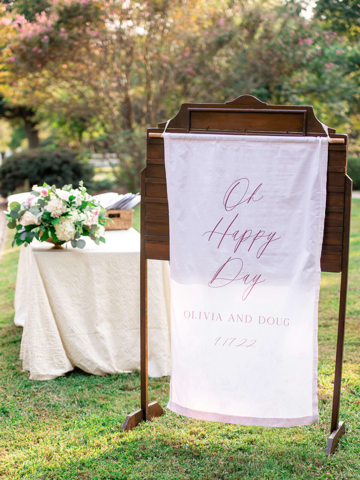 A wooden sign stand displays a white banner with cursive text. In the background, theres a table with a floral arrangement and a book on a grassy lawn.
