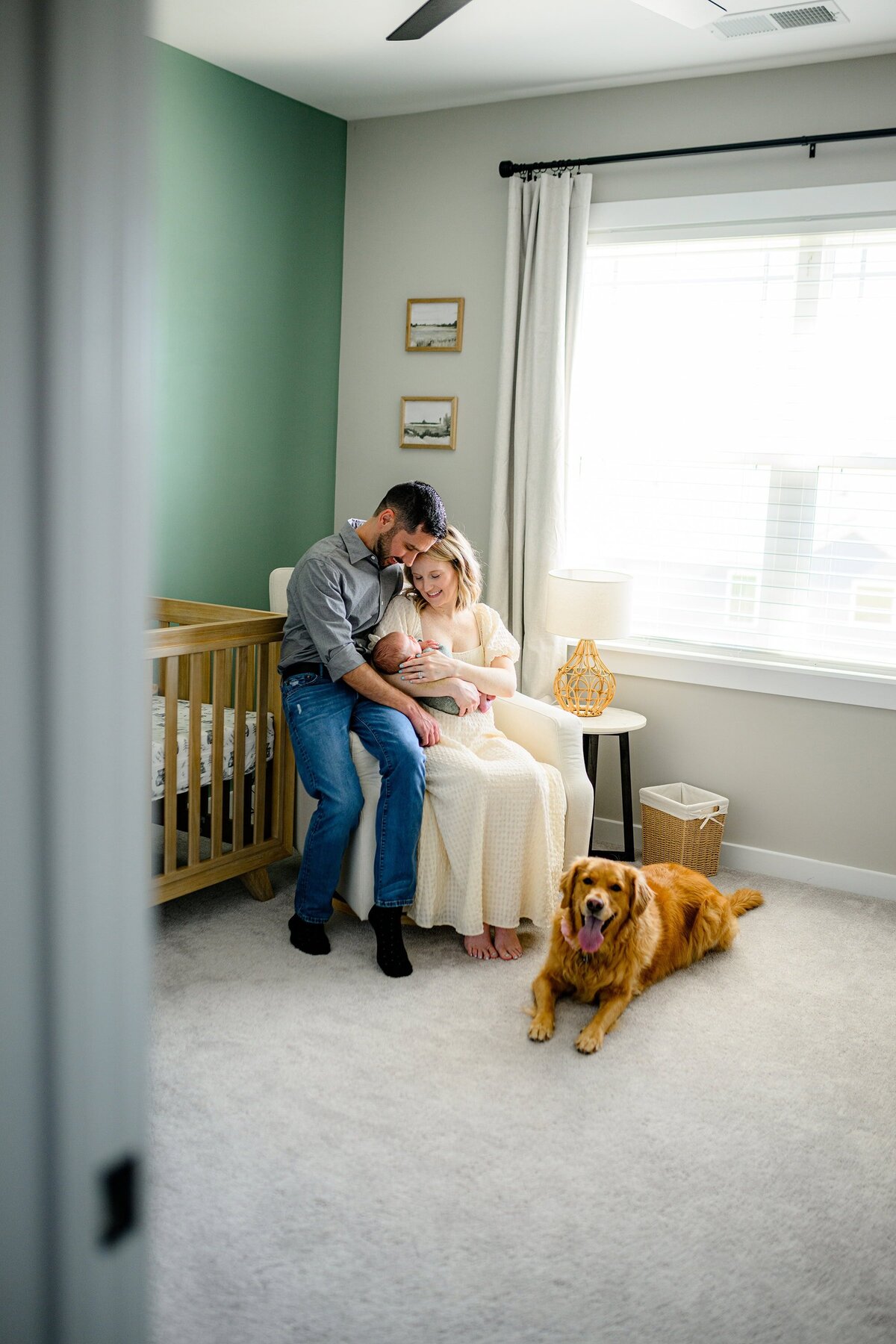 mother and father snuggle thier newborn son while sitting in a chair with their dog beside them