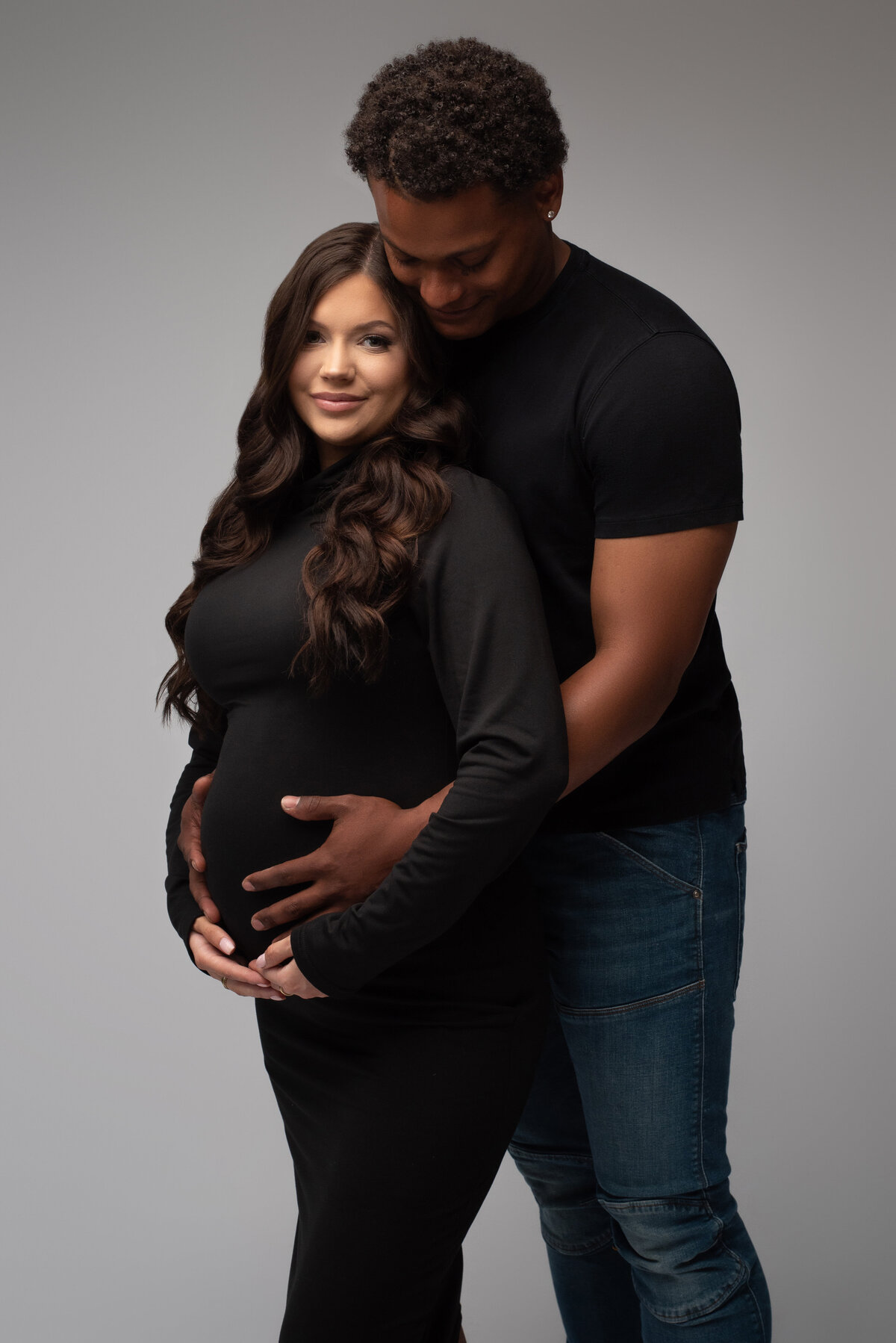 man wearing jeans and black tshirt posing with pregnant woman wearing turtleneck black dress on grey backdrop