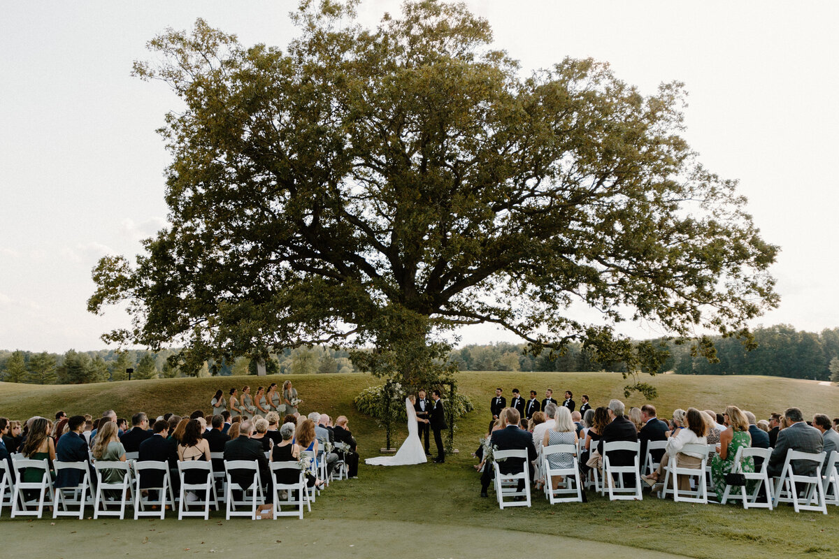 outdoor wedding ceremony taking place under a large oak tree at a golf course