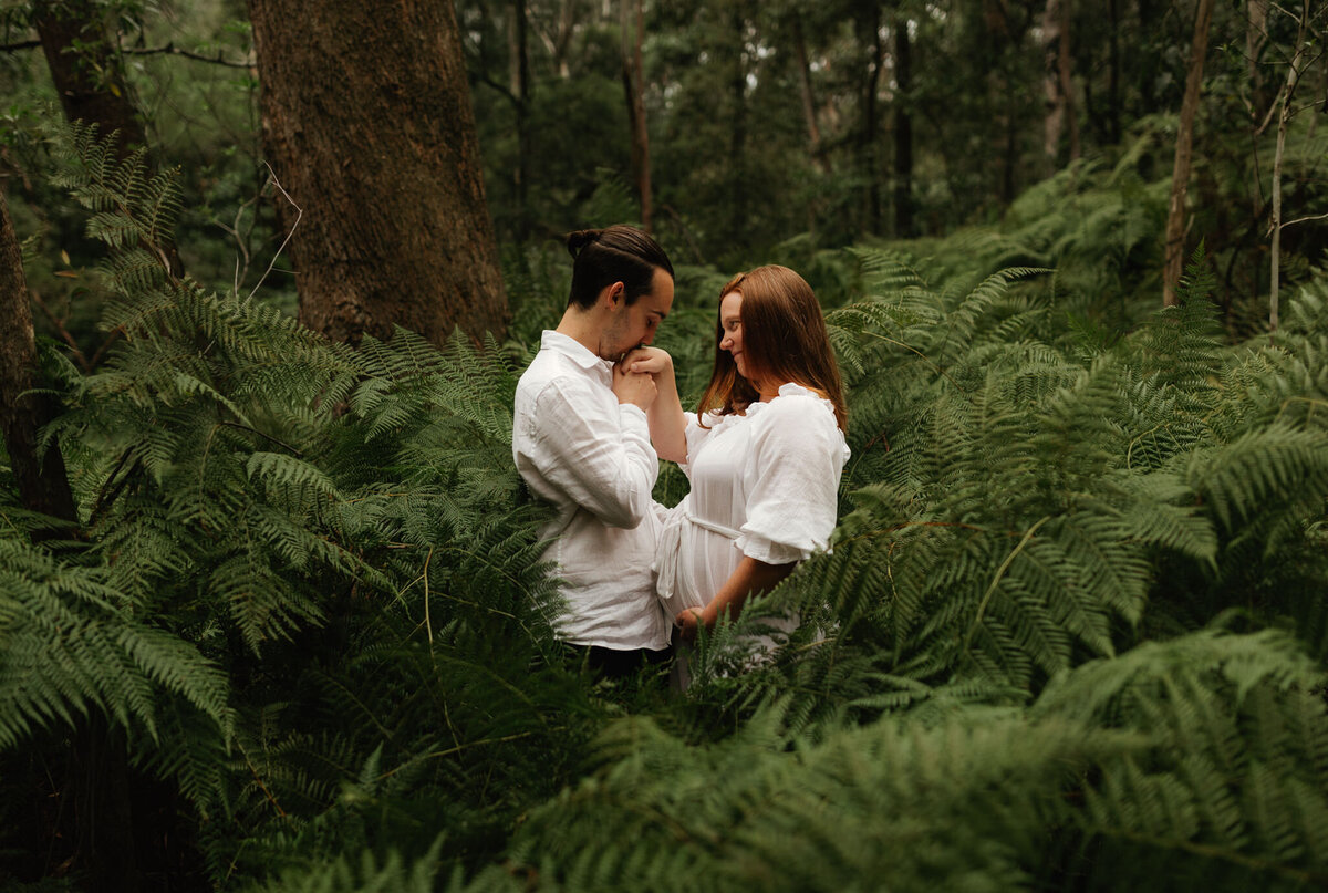 Expectant couple standing close together in a lush forest surrounded by ferns, with the father-to-be gently kissing the mother’s hand in a quiet moment of connection.