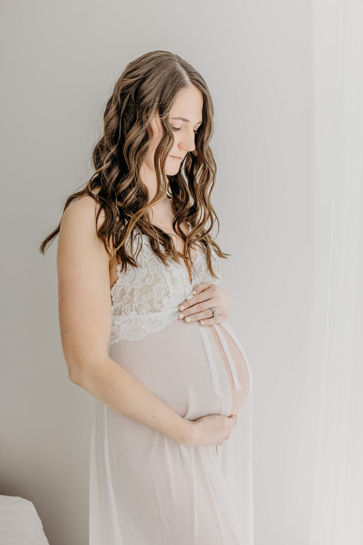 Young mother holding baby in the belly in sheer white intimate maternity photo
