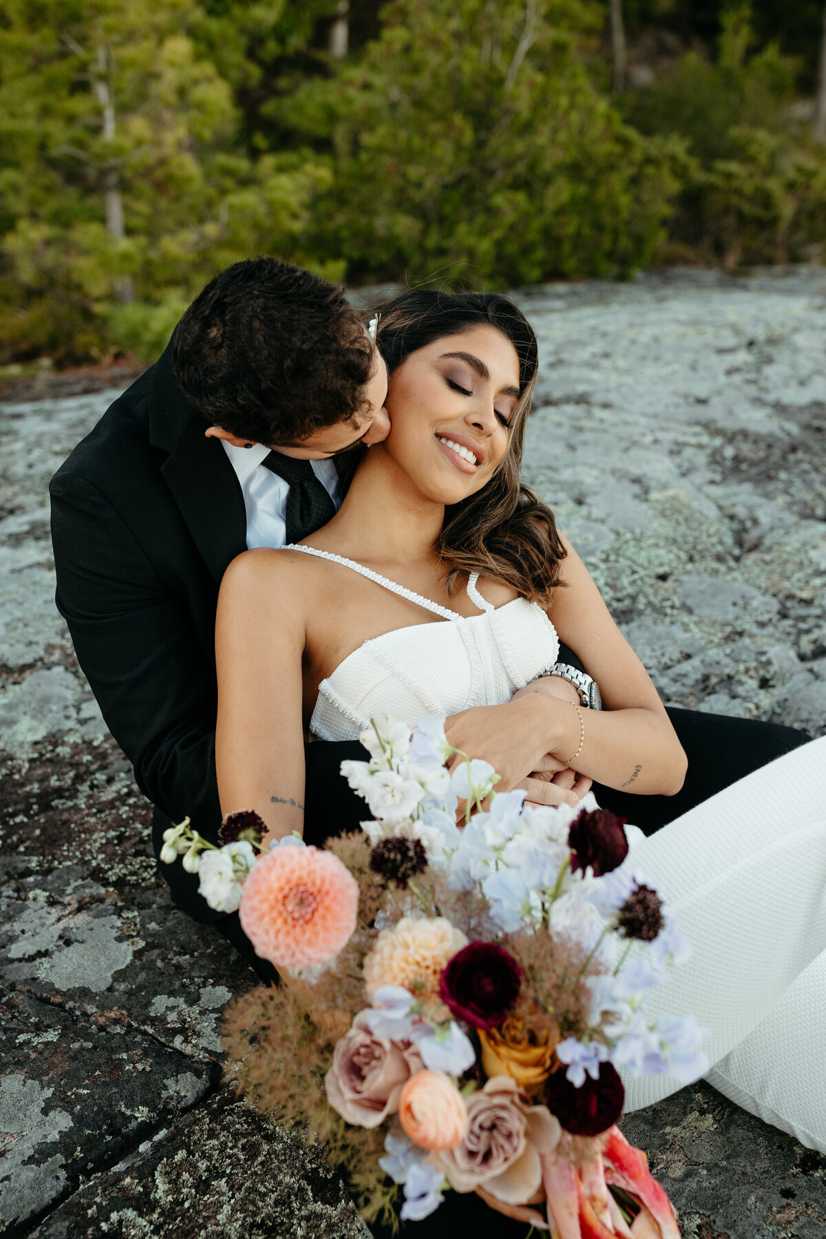 A groom kissing the bride on the cheek as they sit on a rock, with a bouquet and water in the background