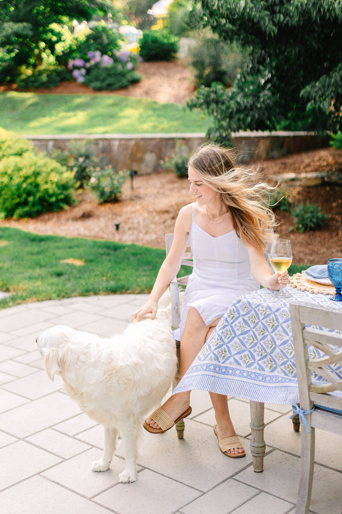 A woman in a white summer dress sits at an outdoor table, petting a fluffy white dog while holding a glass of wine. Her hair flows in the breeze, and the setting features lush greenery and a beautifully set table with blue and white decor.
