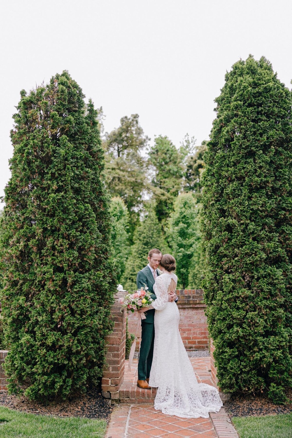 A couple embraces surrounded by lush greenery and tall hedges.