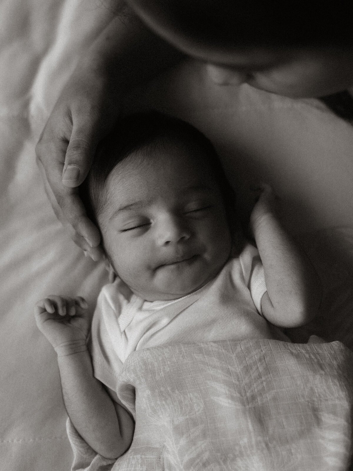 A black and white photo of a newborn baby smiling softly while lying in bed, with a parent’s hand gently cupping their head.