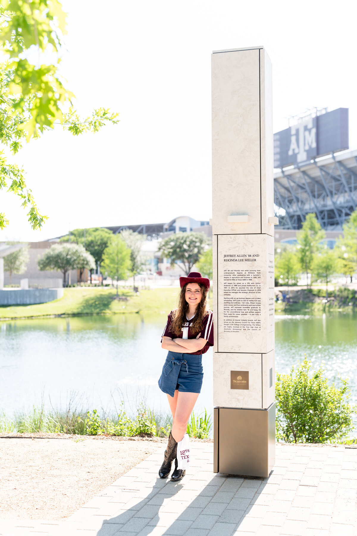 Texas A&M senior girl leaning against column in Aggie Park with Kyle Field in background while wearing Aggie jersey, maroon cowboy hat and jean skirt
