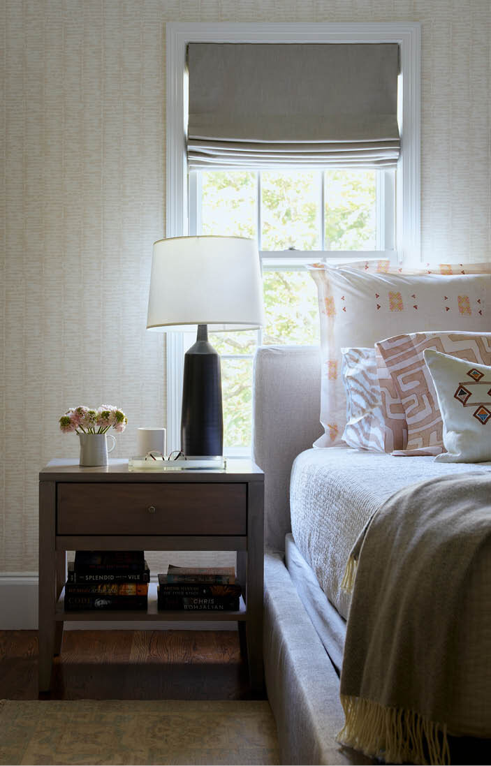 View the primary bedroom in a historic Hingham home, featuring a soft grey wood bedside table, black ceramic lamp, custom upholstered bed, tone-on-tone wallpaper, and subtle soft bedding patterns.