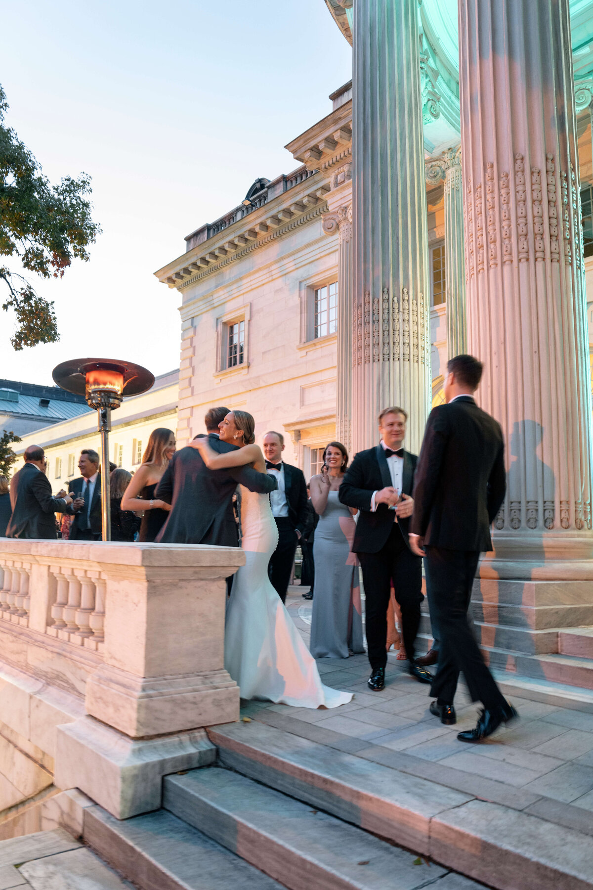 A group of elegantly dressed people gathers outside a grand building during an evening event. A couple embraces at the forefront, with others standing nearby, chatting and smiling. The setting includes large columns and soft lighting.