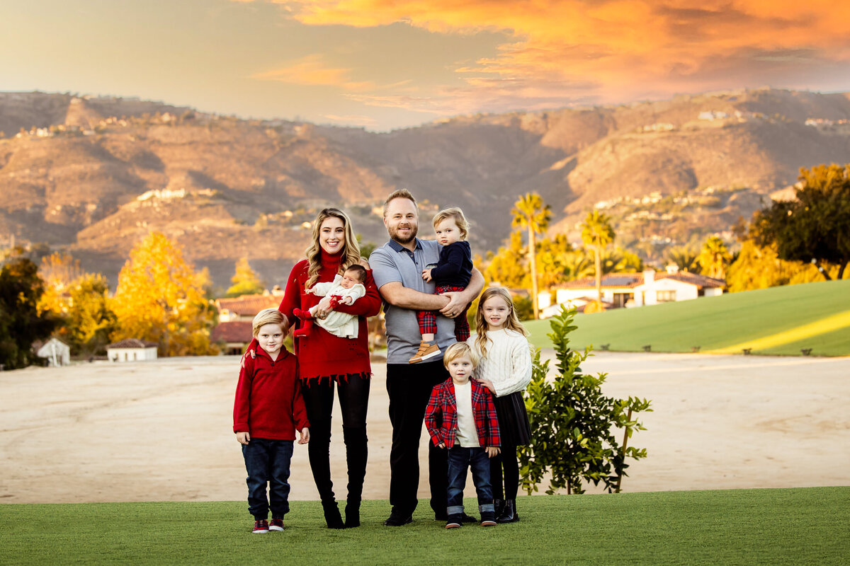 portrait if a family of 7. they are dressed in red and white. they are standing on green grass with a view if the hills at sunset behind them