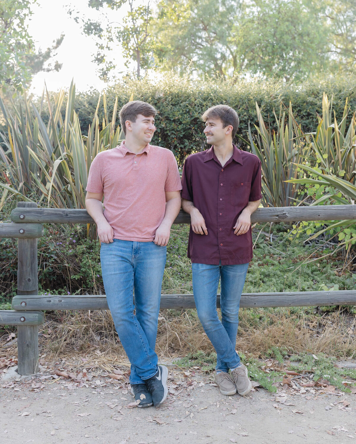 Two adult brothers with dirty blonde hair wearing blue jeans and different shades of red, leaning their elbows on the top rung of a wooden park fence, looking at each other sideways and smiling.