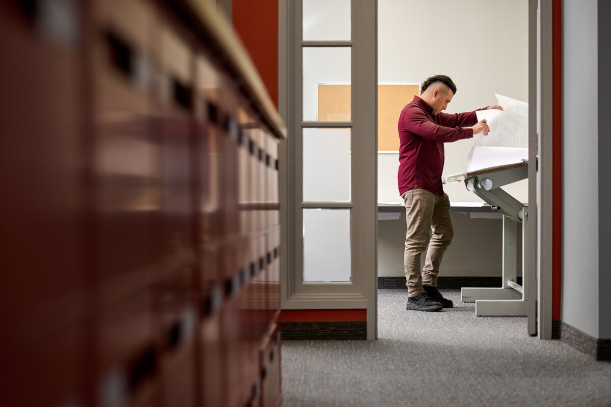 An engineer at Brumar Engineering in Ontario reviewing architectural blueprints at a drafting table. The office setting highlights a balance of traditional and modern engineering practices, captured by Brandon Marsh Photo & Video Productions.