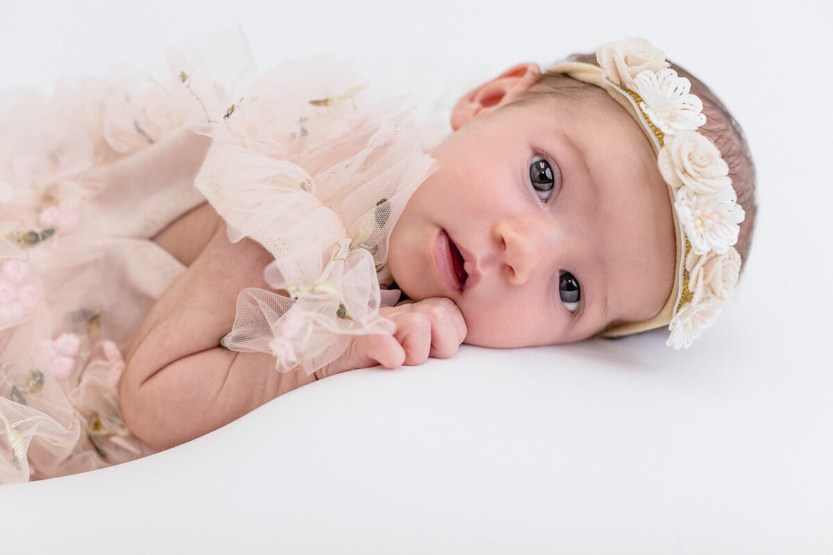 A baby wearing a floral headband and a frilly, light pink outfit lies on a white surface, gazing curiously upward. The soft textures and pastel colors create a serene and gentle atmosphere.