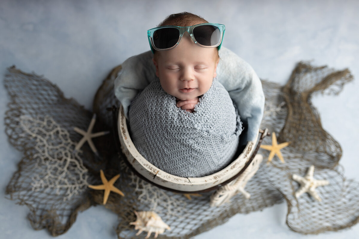 Smiling newborn baby with teal sunglasses wrapped in grey knit blanket surrounded by starfish and netting by Dallas newborn photographer.