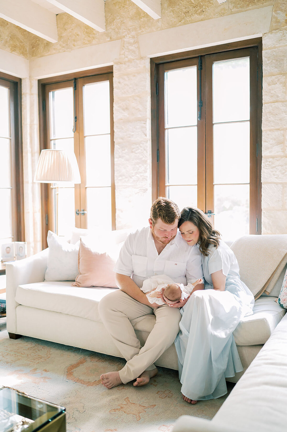 During an in-home lifestyle newborn photo session with Austin Newborn Photographer Teressa Jane, a mother and father sit together on their couch, lovingly holding their newborn. Their faces glow with warmth and joy as they embrace their new baby in the comfort of their home