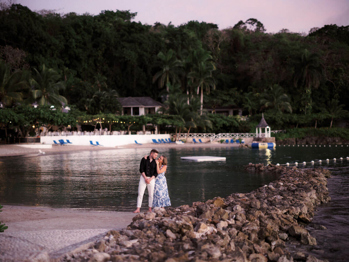 The engaged couple is standing on the seashore in front of ocean, resort and many trees at Roundhill Hotel and Villas, Jamaica.