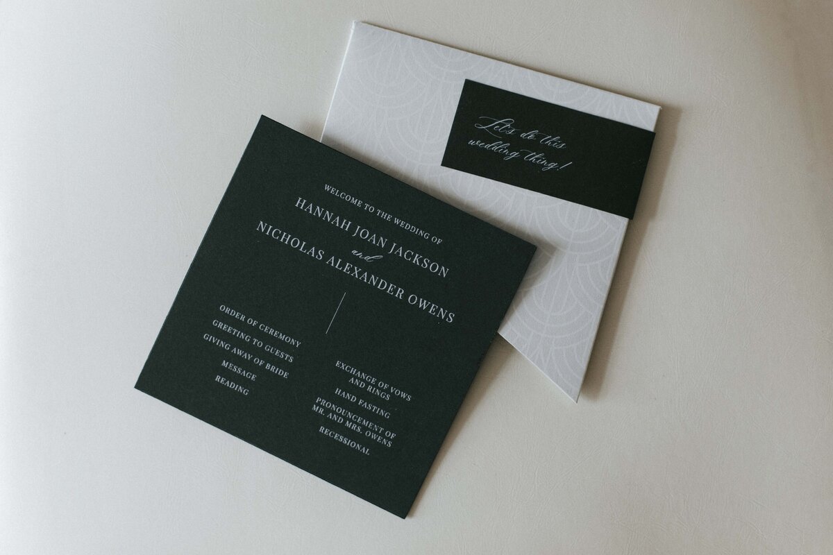 Dark green wedding invitation with white cursive font atop ivory and white textured envelope.