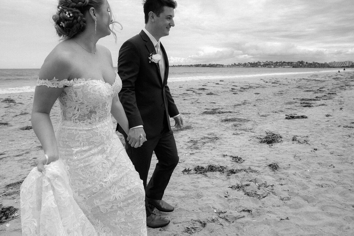 Bride laughing while walking barefoot through sand on the shore of Boston, MA, holding up her dress.