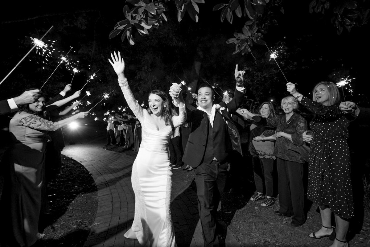 Black and white photograph of a bride and groom celebrating with their hands in the air, smiling as they run through their sparkler exit at The White Home in Rock Hill, SC.