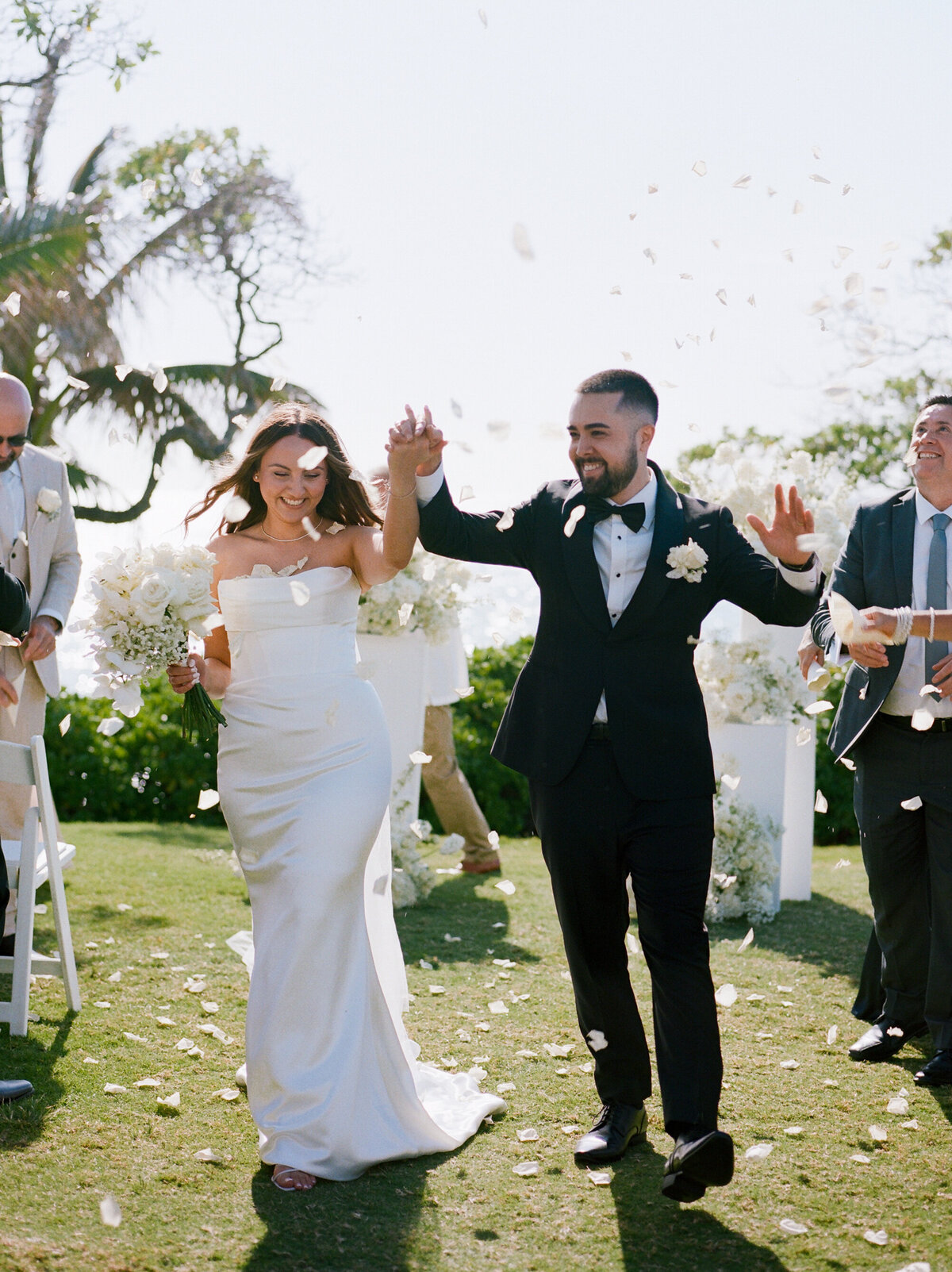 A joyful couple walking down the aisle in Hawaii as petals fall around them, smiling and holding hands—captured on nostalgic film by Kate Winters Photography