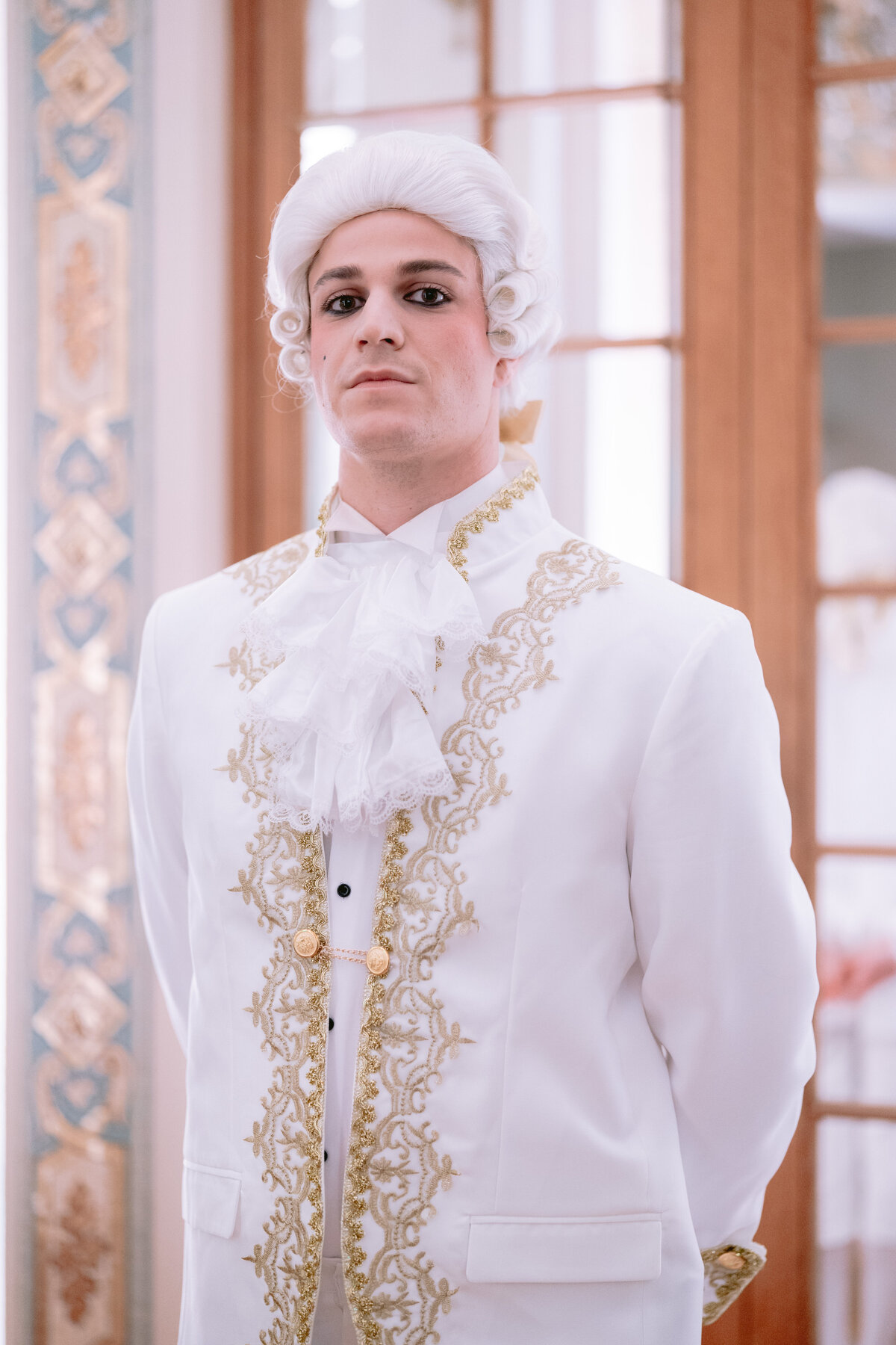 The photo was taken at the luxurious Shangri La Hotel & Ritz Paris Hotel in Paris, France, by the talented photographer Matteo Coltro, with videography by Zen Film Works. In the image, a person in an elaborate historical outfit stands confidently, exuding elegance and charm. They are adorned in a white wig with cascading curls, a white suit embellished with intricate gold embroidery, and a stylish ruffled cravat. The background showcases ornate decor and a window with beautiful wooden framing, adding to the overall grandeur of the scene.