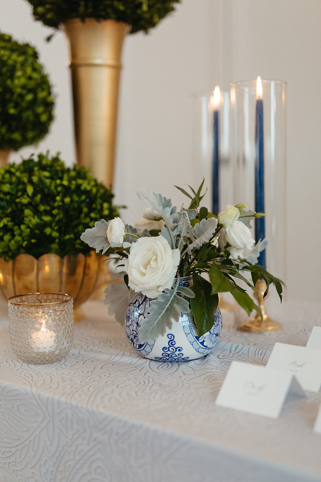 Flower arrangement in blue and white vase with candles and white place cards atop and white linen table.