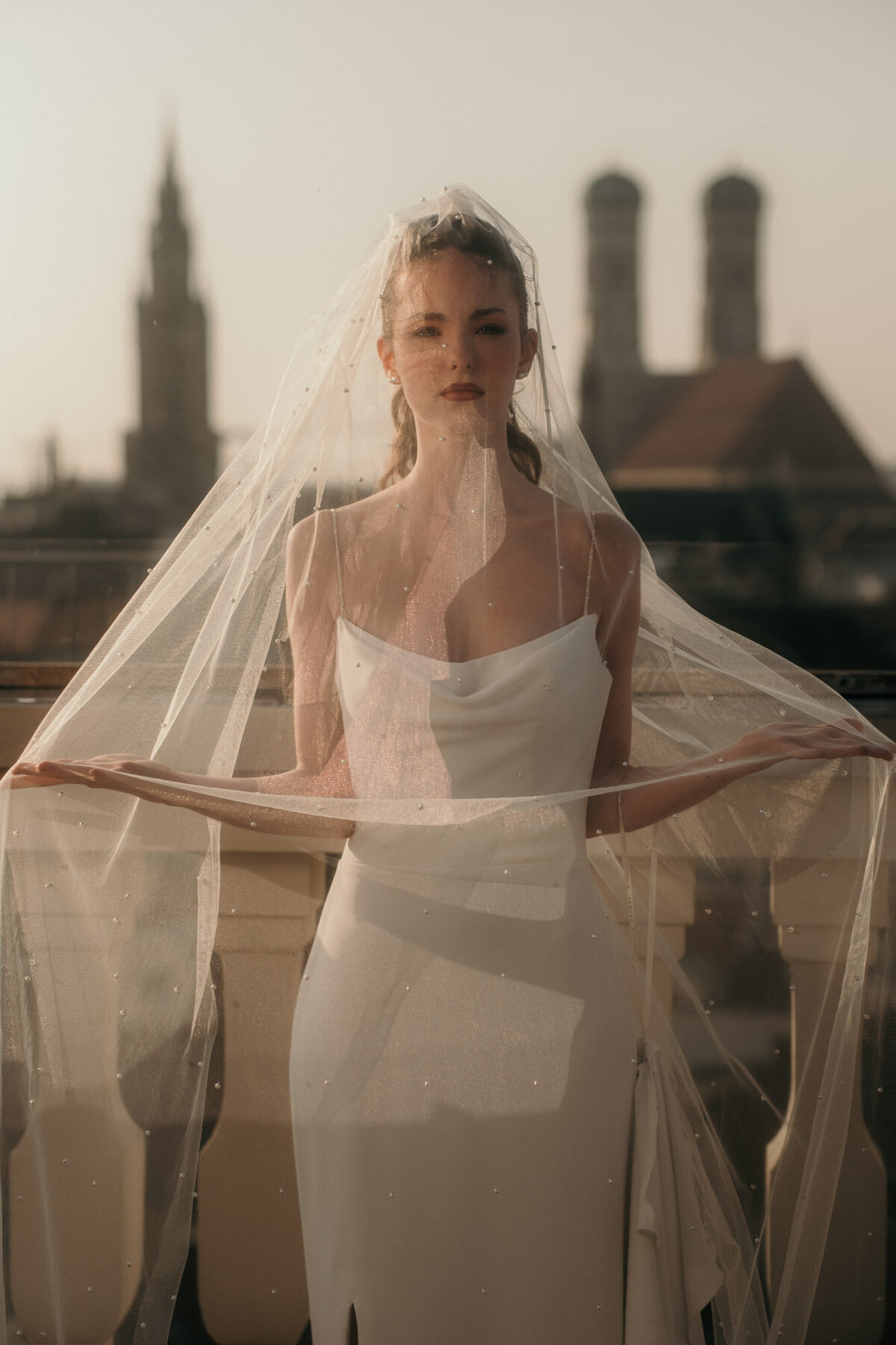 Mit der Münchener Innenstadt im Hintergrund hält das Brautmodel den Schleier mit flachen Händen hoch.
