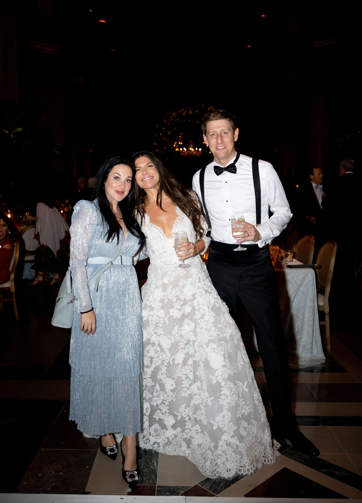 Three people are posing and smiling indoors at what appears to be a formal event. The woman on the left is wearing a light blue dress, the woman in the center is in a white lace wedding gown, and the man on the right is in a tuxedo. They are standing close together.