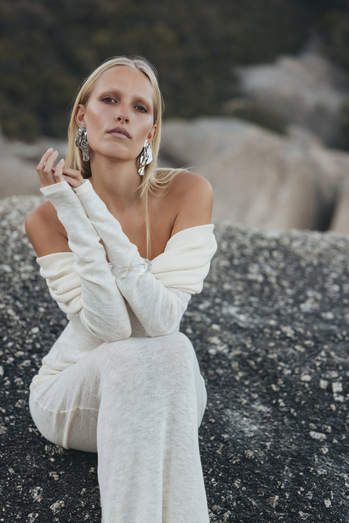 Model Johanna poseert zelfverzekerd in een witte jurk op een rots in Kaapstad, met zilveren oorbellen en een serene blik.