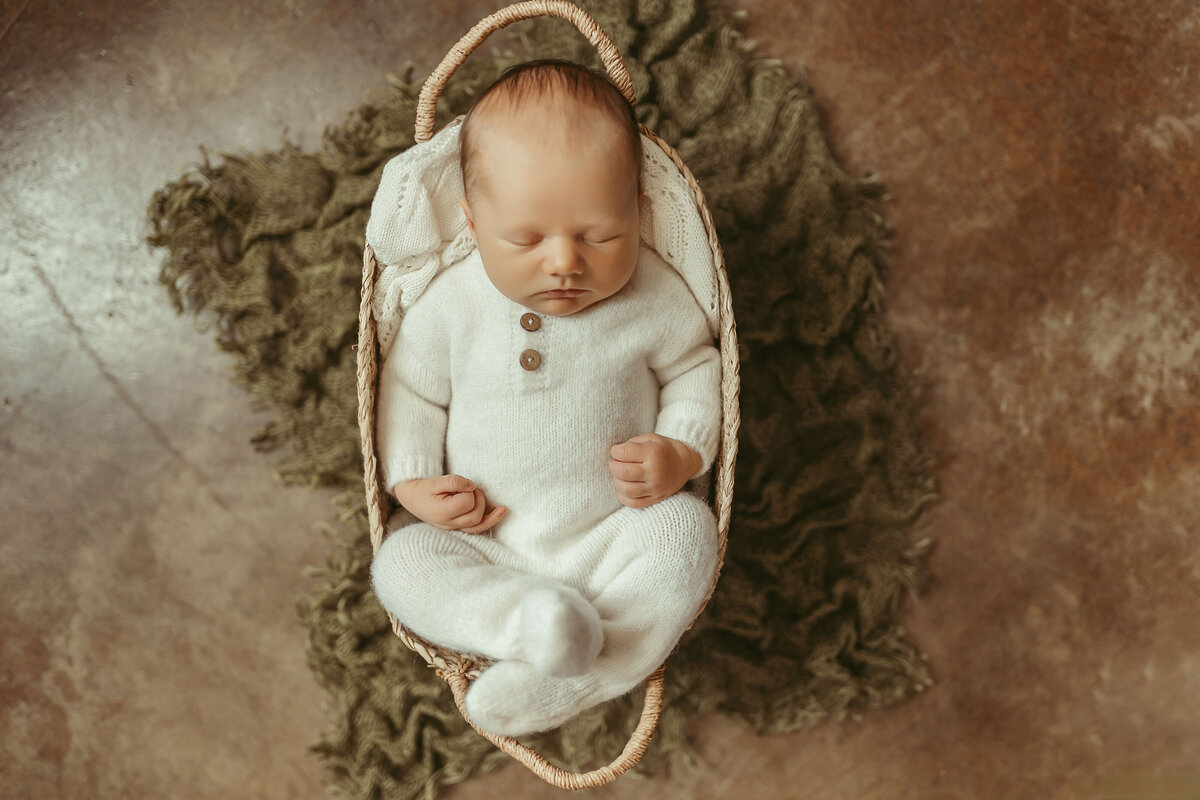 baby wearing a cozy white onesie with buttons. He is sleeping inside a basket with his fists closed and eye shut