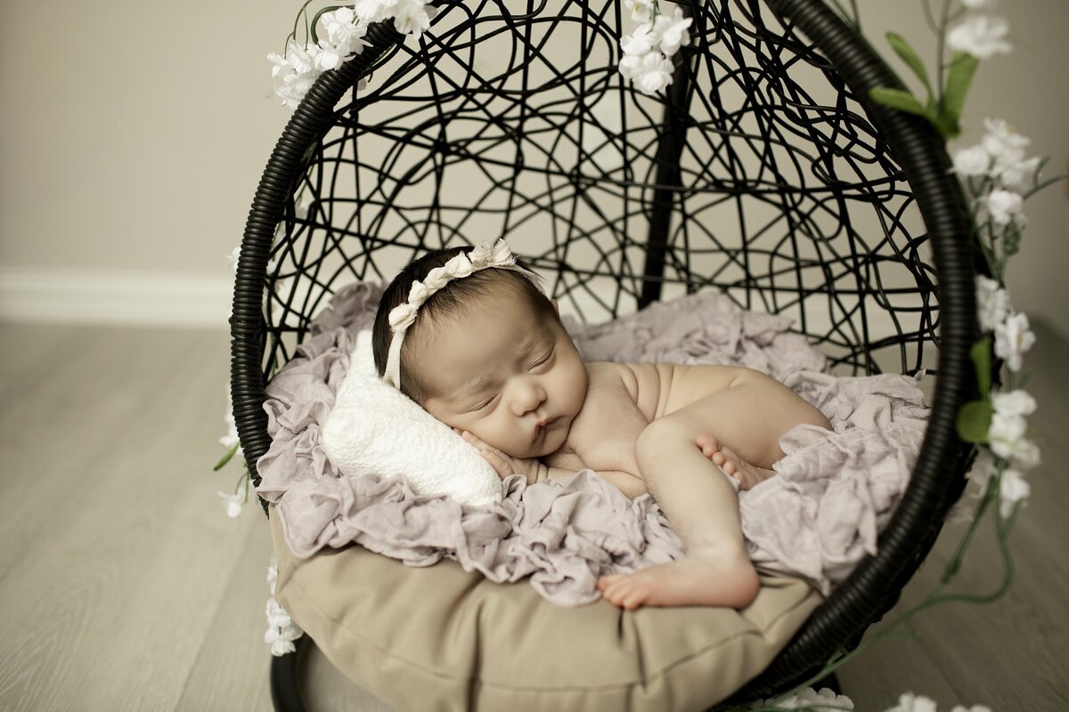Newborn baby girl peacefully resting in a black wicker hanging basket, nestled in soft blankets, creating a cozy and artistic portrait