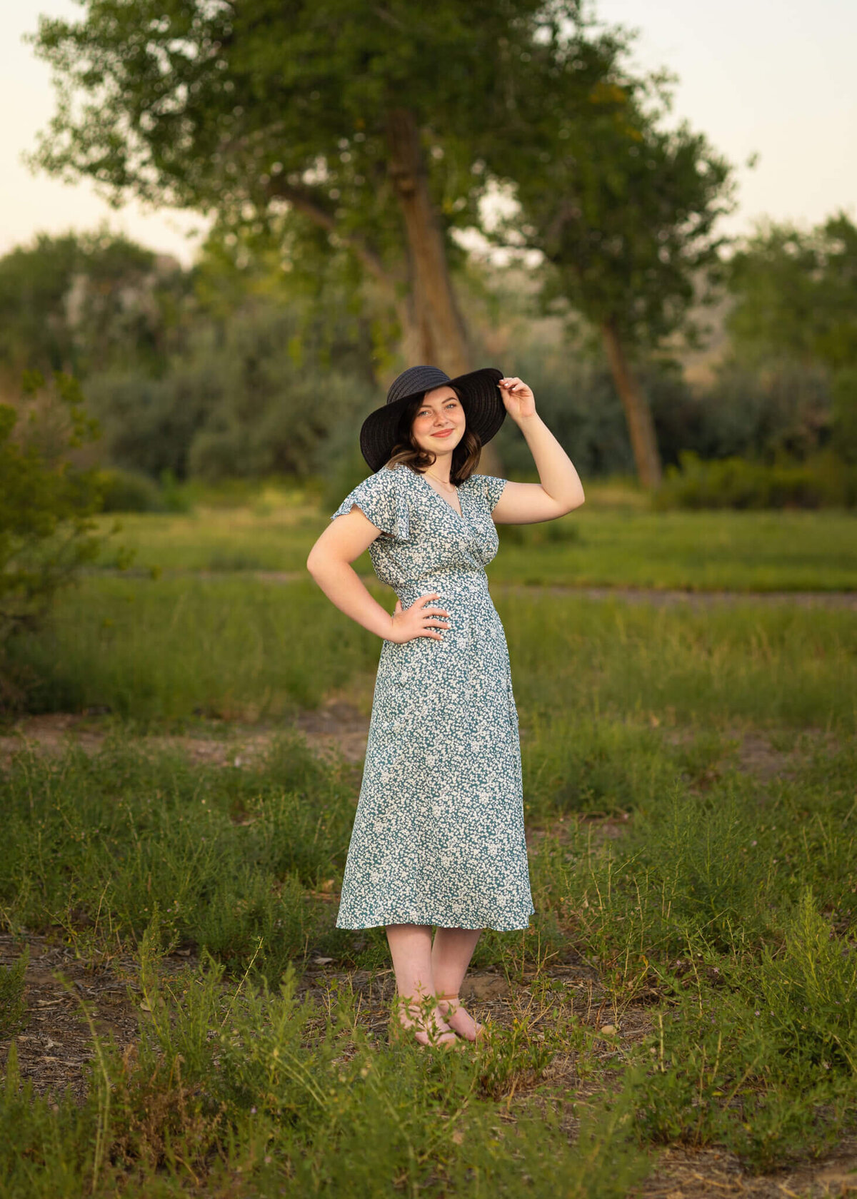 high school senior girl in green floral dress with a black floppy hat standing in a green field