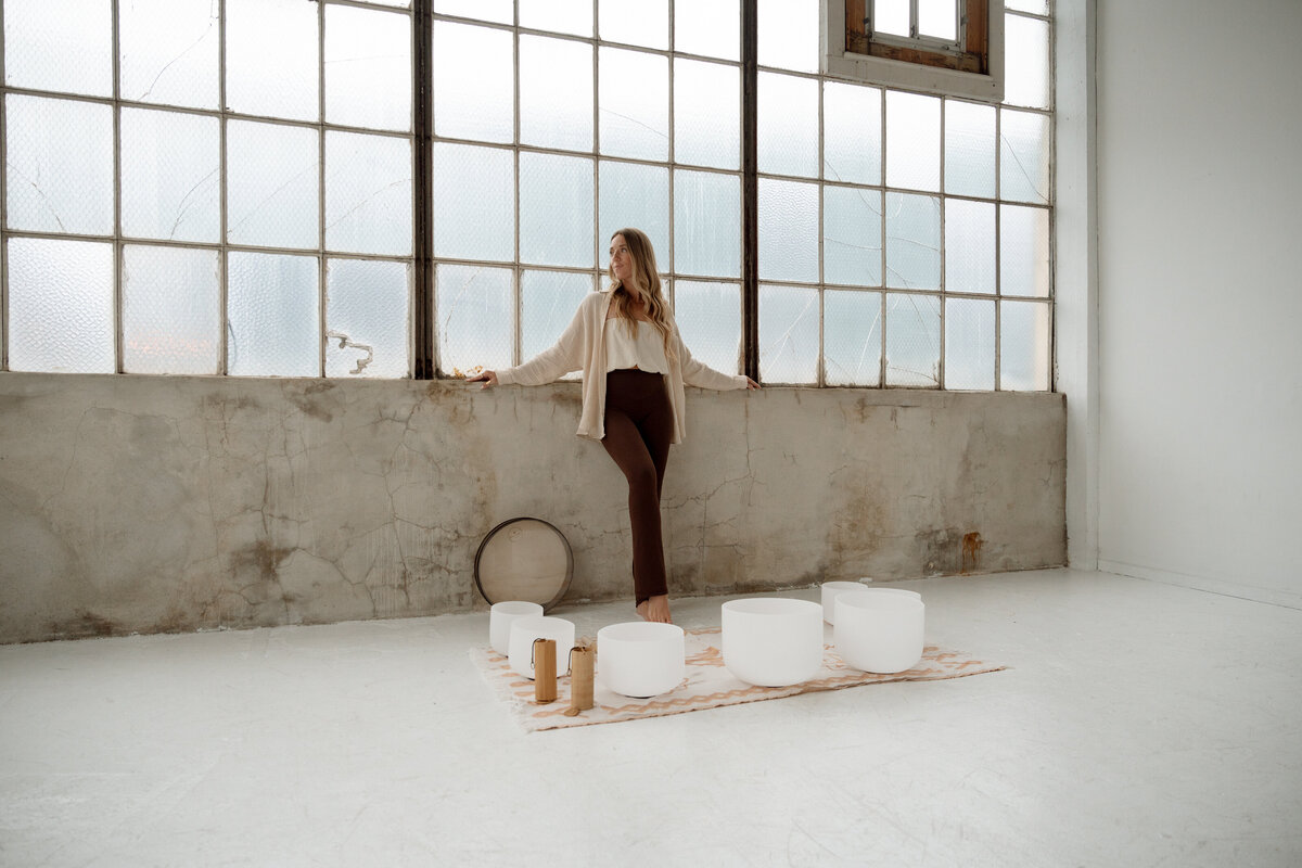 Creative branding session in Irvine featuring a woman standing by tall industrial windows with sound bowls arranged on the floor.