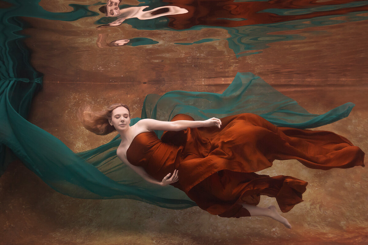 A mesmerizing underwater portrait by Lindsay Walden Photography featuring a woman gracefully floating in a rich, rust-colored gown with emerald fabric billowing around her. The soft golden tones of the backdrop and serene expression on her face create a timeless, elegant atmosphere, highlighting the artistry of fine art underwater maternity photography.