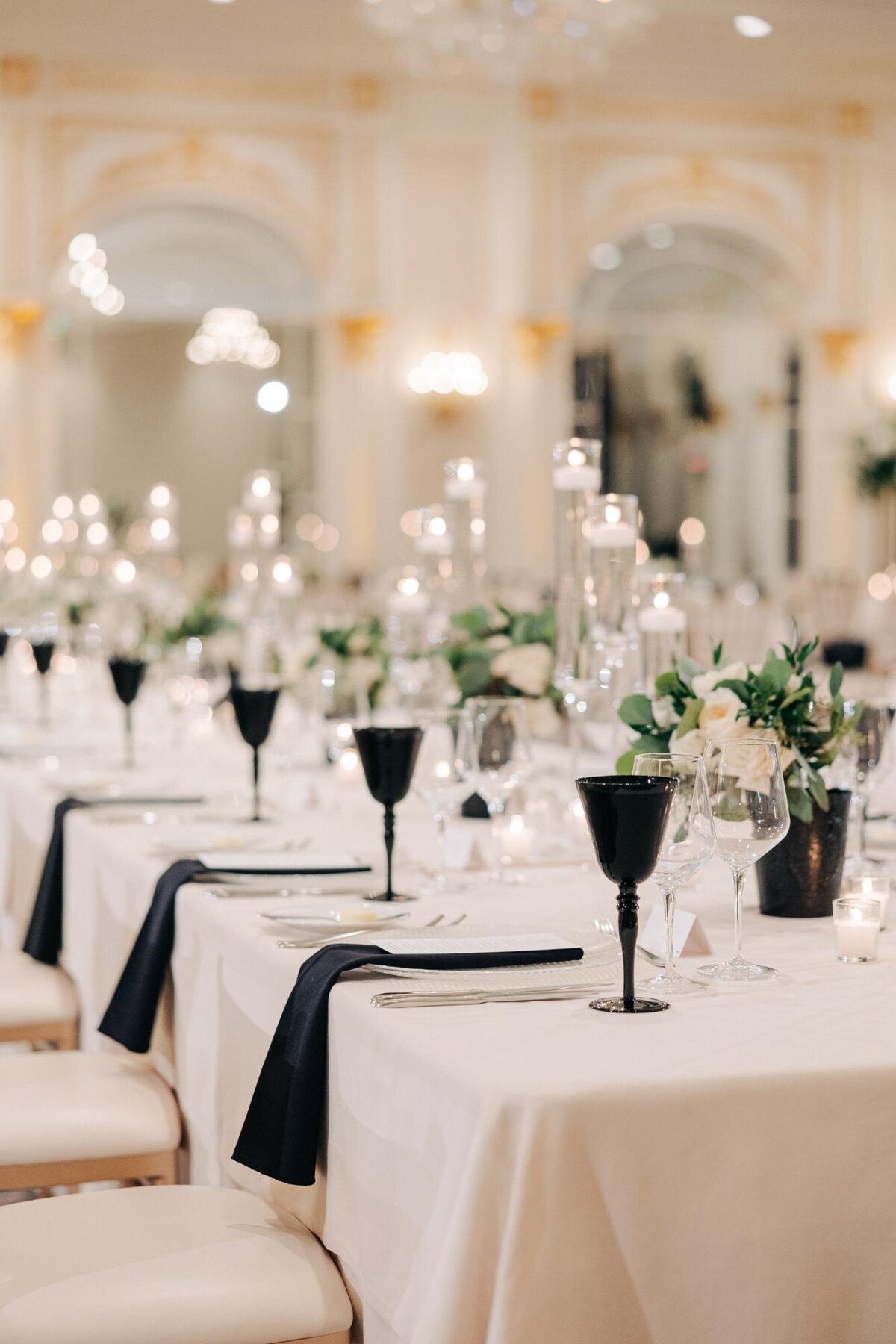 Elegant dining setup with white linens, black glassware, and floral centerpieces in a sophisticated ballroom.