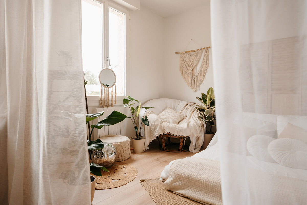 Ambiance cocooning pour ce studio photo en Vendée de Laura. Décor composé d'un lit, d'un grand fauteuil rond recouvert de plaid, de plantes exotiques et de voilages blancs.