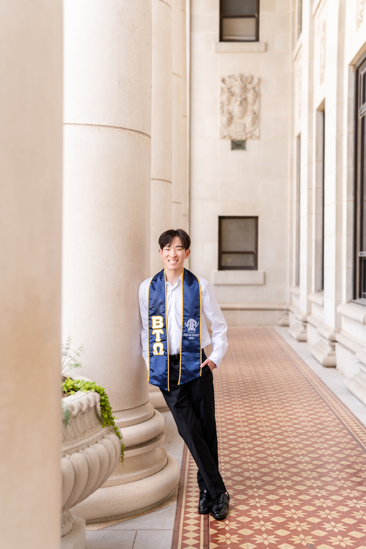 Texas A&M senior guy wearing stole and leaning against column with hands in pocket and smiling at the Administration Building