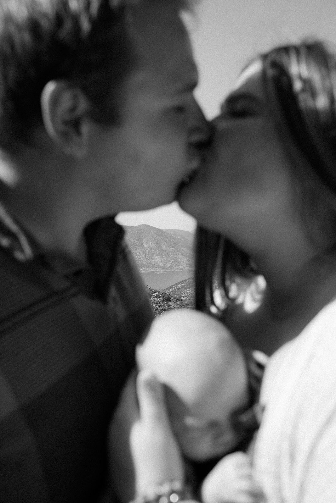 Mother and father kiss over infants head as camera focuses beyond them to a nearby lake. 