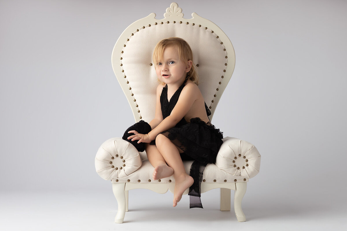 Toddler sitting on a vintage chair holding a black stuffed animal, taken by family photographer in Dallas.