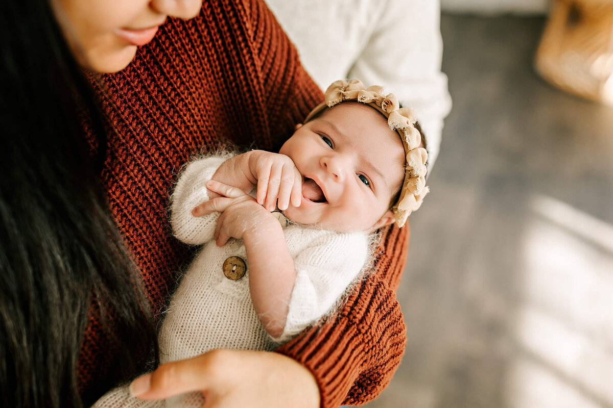 A person in a brown sweater cradles a smiling baby wearing a knitted white outfit and a floral headband. The baby is holding their hand to their mouth. The background is softly blurred, highlighting the gentle interaction.