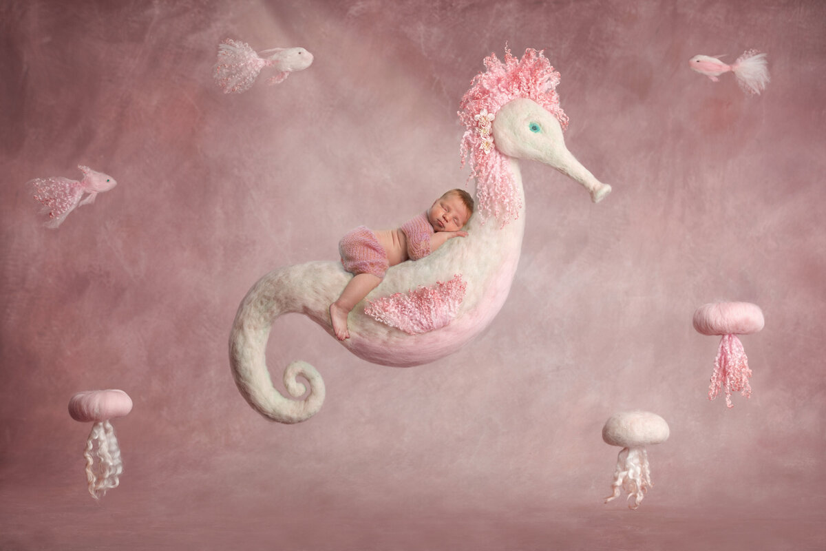 Ginger newborn girl floating on a wool seahorse suspended in the air.  There are also pink and white wool felted jellyfish.  Baby girl is sleeping.  Composite image.