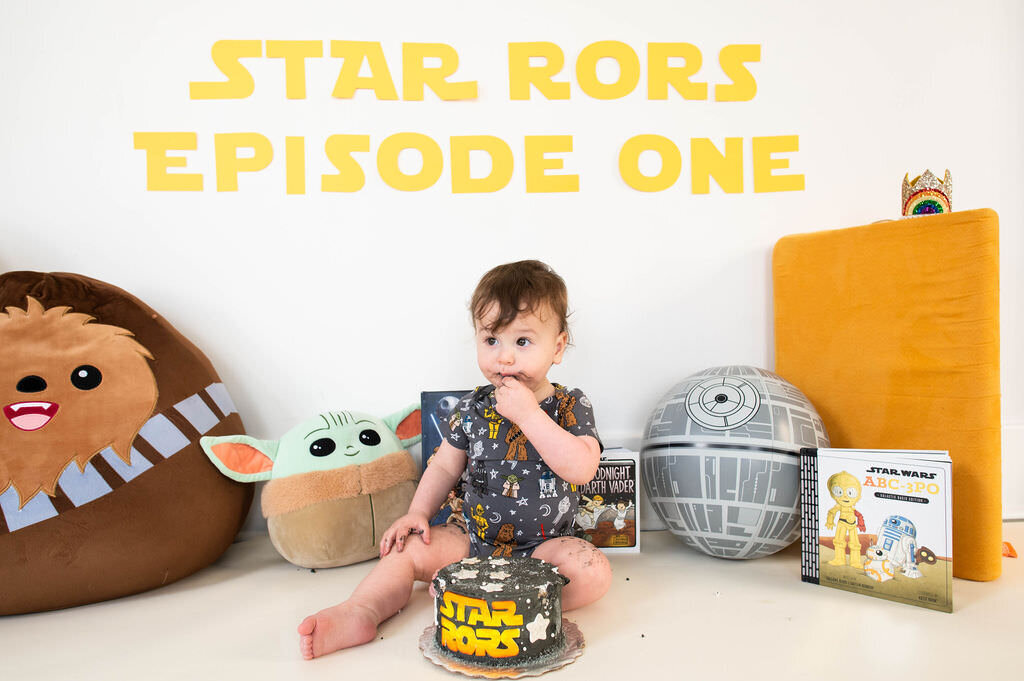 A baby sitting on the ground with Star Wars toys around him and a cake in front of him.
