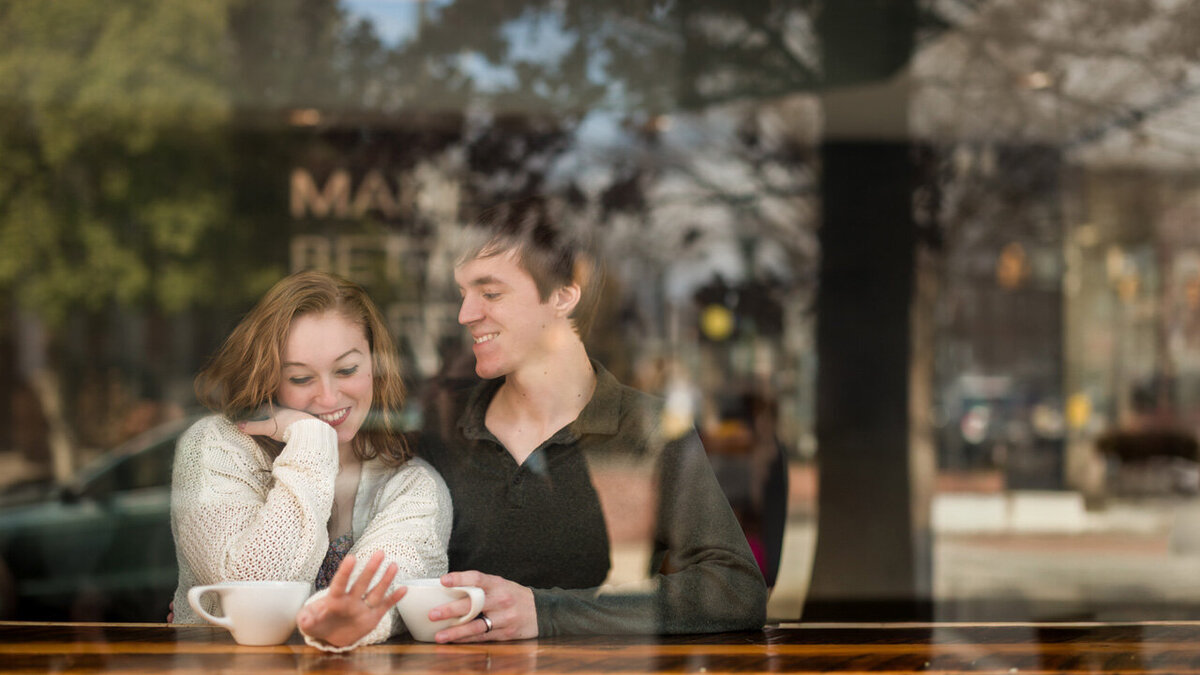 Engaged couple in a cozy coffee shop admiring a sparkling engagement ring during their engagement session, showcasing an intimate and romantic moment in an urban setting.