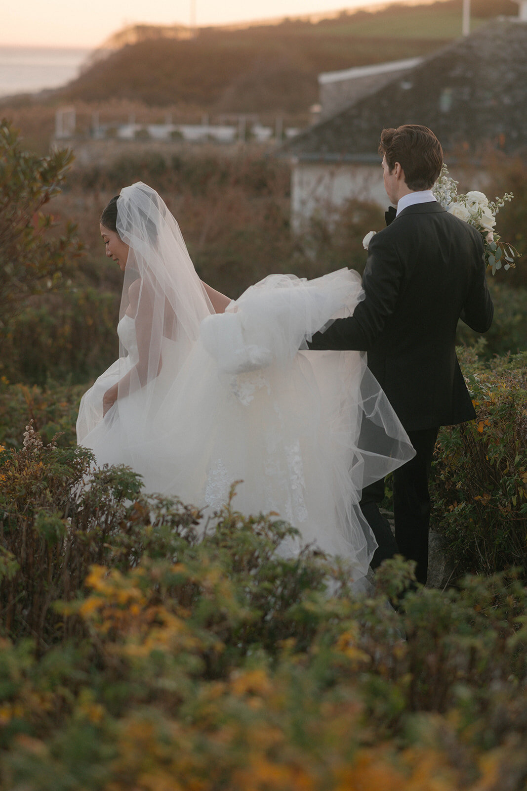 Editorial wedding moment of a bride in a Vera Wang gown walking with her groom through coastal greenery at the iconic Ocean House in Watch Hill, Rhode Island.