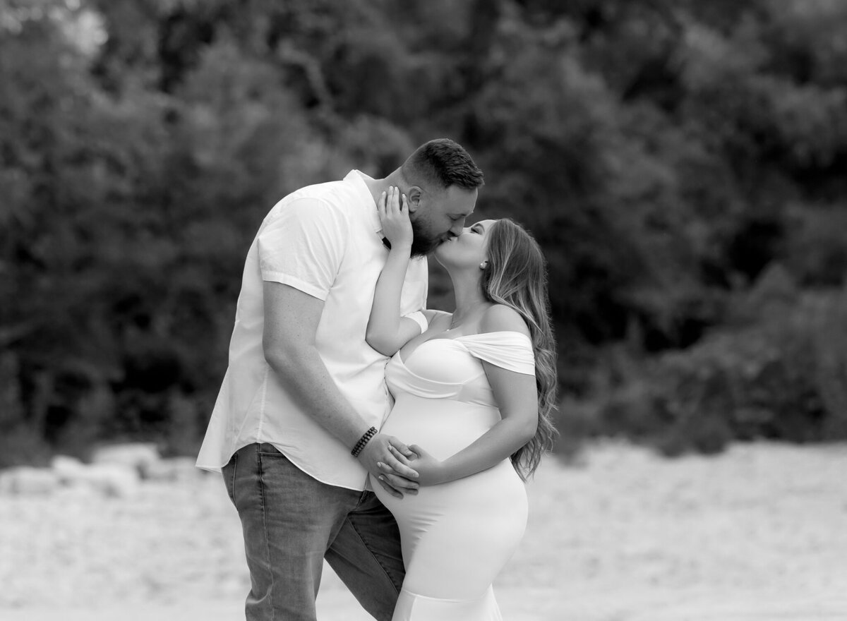 Black and white outdoor maternity portrait of a couple kissing, with the expecting mother in a form-fitting white dress in Westlake.